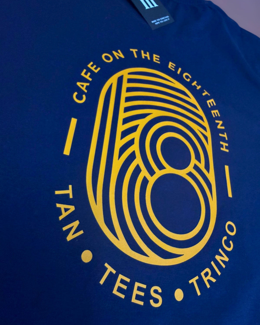 A close-up of a royal blue t-shirt featuring a large yellow graphic print. The design displays a stylized number 8 integrated with a fingerprint pattern, surrounded by the text 'CAFE ON THE EIGHTEENTH', 'TAN', 'TEES', and 'TRINCO'.