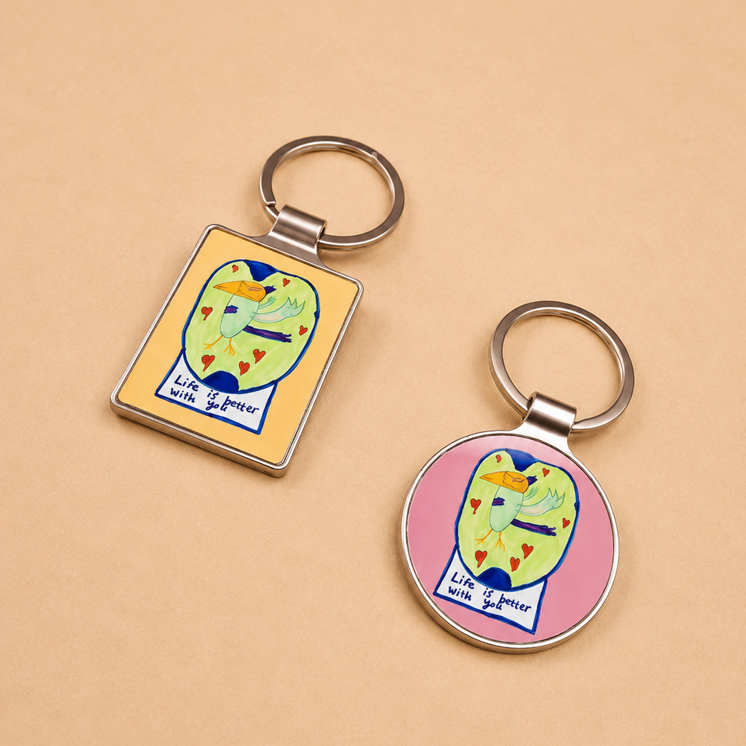 Two metal keychains with colorful cartoon cat illustrations on a warm beige background. One keychain has a rectangular yellow face and the other has a round pink face, both with silver key rings.
