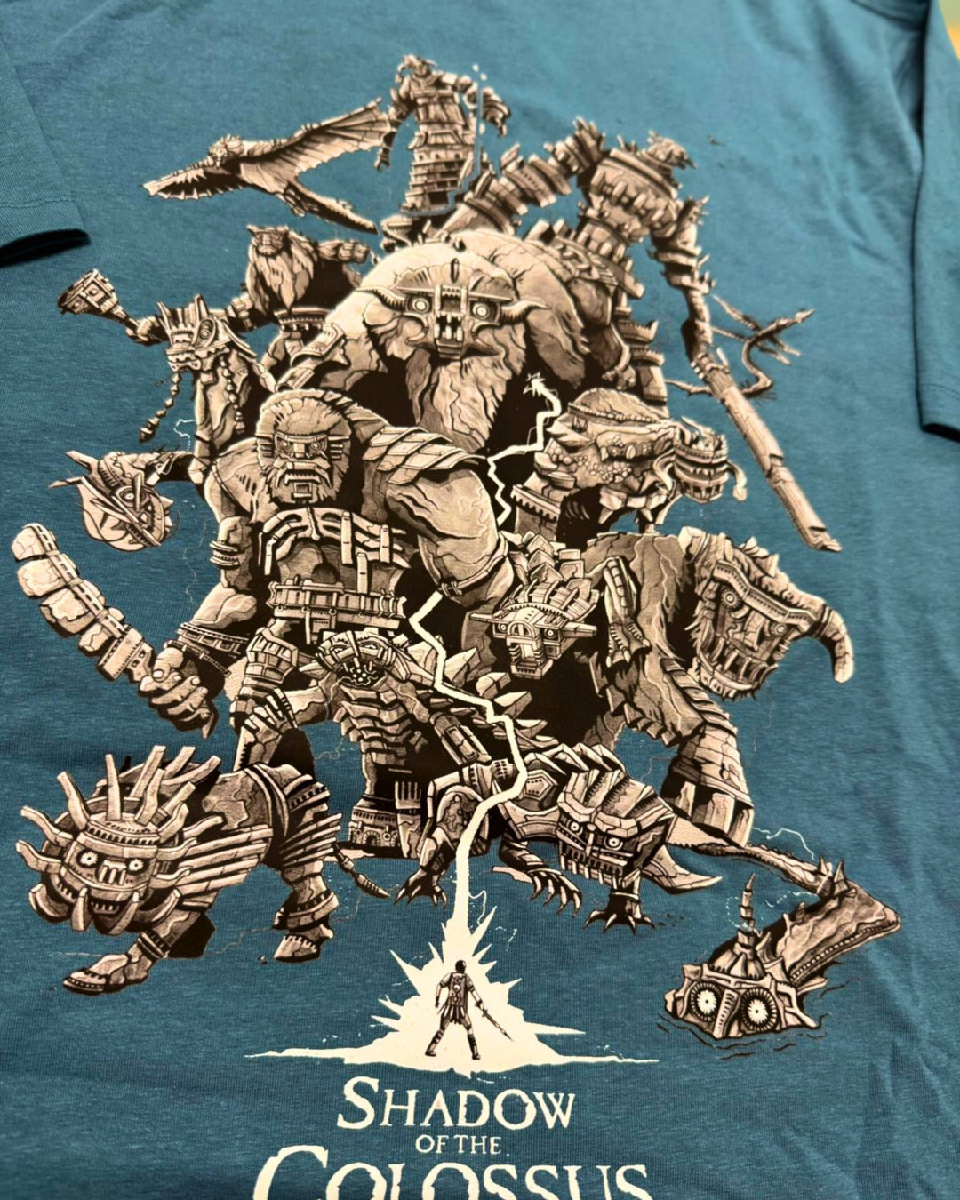A close-up of a teal-blue T-shirt featuring a large beige graphic print of multiple fantasy colossus creatures and the text 'Shadow of the Colossus' near the bottom.