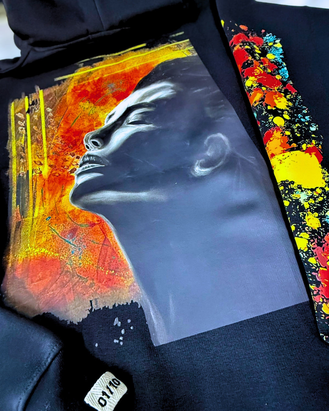 A close-up of a black hoodie featuring a large graphic print of a face in profile against a fiery orange, yellow, and red abstract background. A second colorful abstract print is visible on the sleeve.