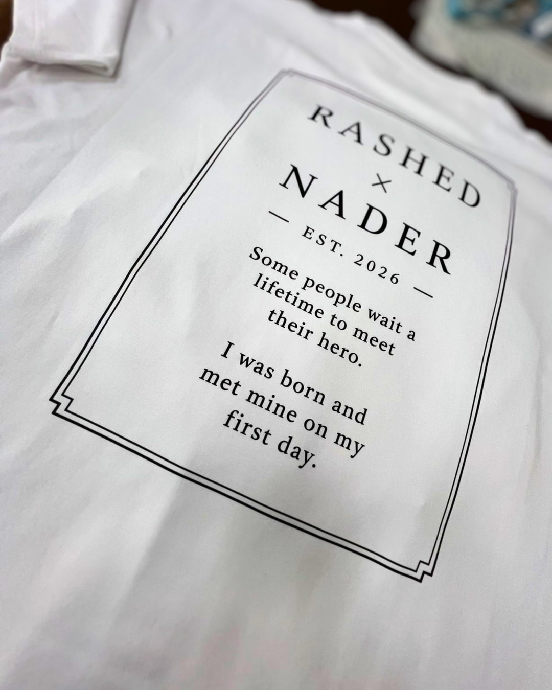A close-up of a white T-shirt with a printed black graphic on the front. The design features the text 'RASHED x NADER EST.