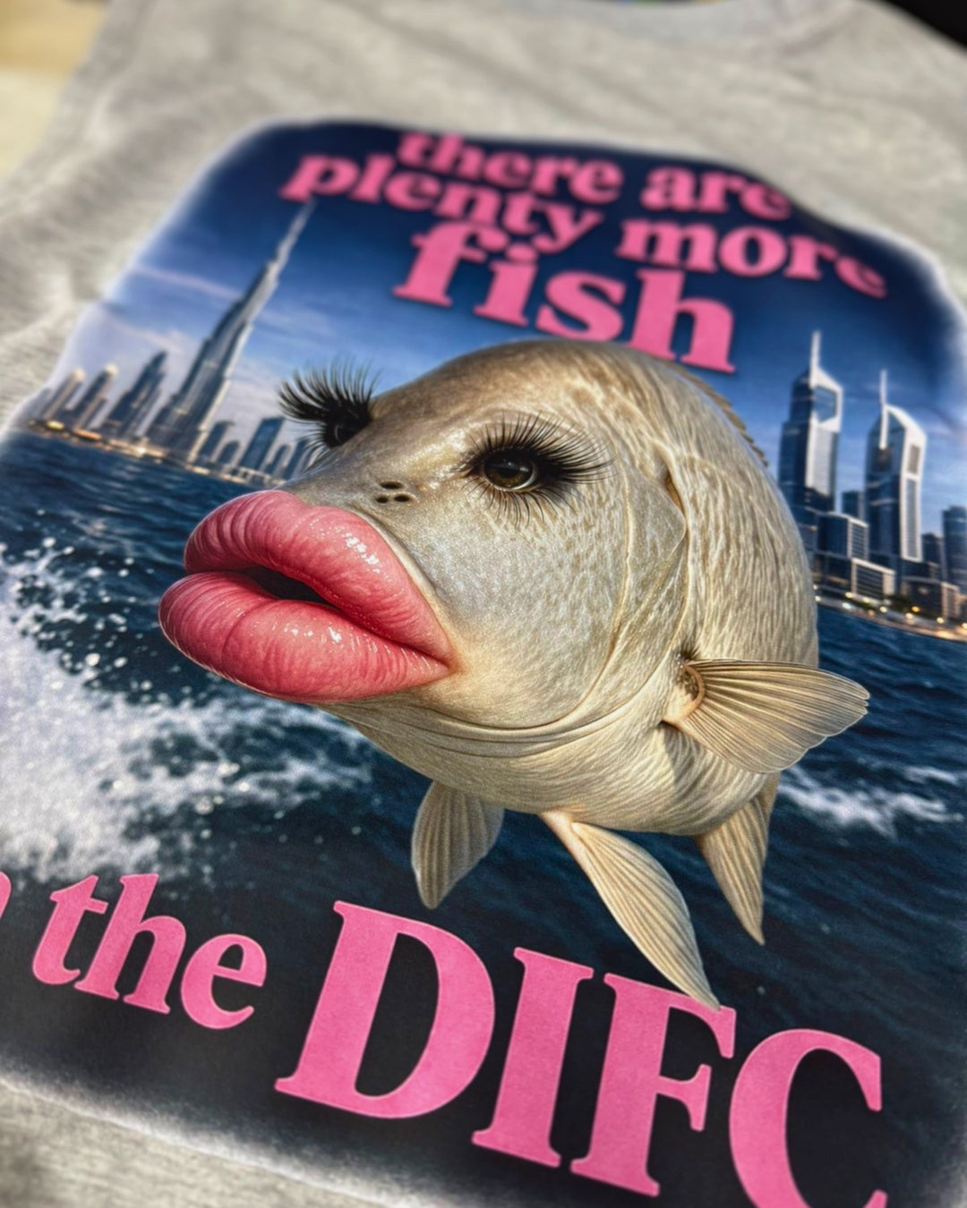 A close-up of a gray heather t-shirt featuring a graphic print of a female-looking bass fish with eyelashes and pink lips, set against a city skyline and blue water, with bold pink text reading 'there are plenty more fish' and 'the DIFC'.