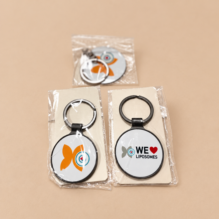 Three oval keychains in clear plastic packaging on a beige background. Two keychains are displayed on cream card inserts, and one is shown in its packaging in the back.