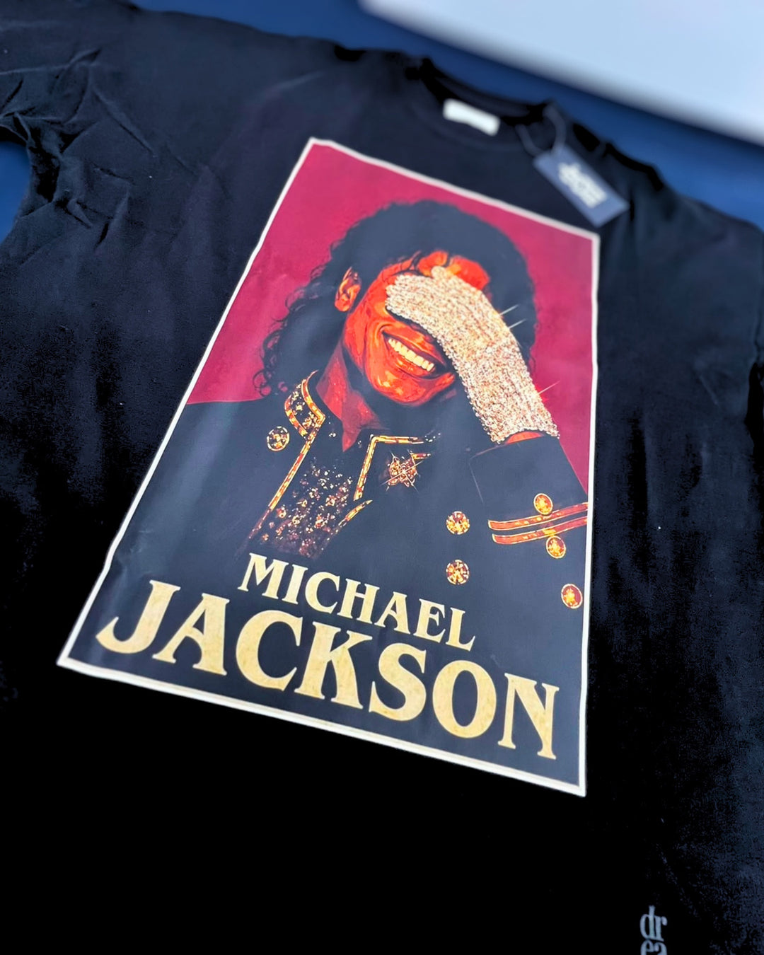 A black short-sleeve graphic T-shirt laid flat, featuring a large rectangular print of Michael Jackson with gold text reading 'MICHAEL JACKSON' and a red background in the print.