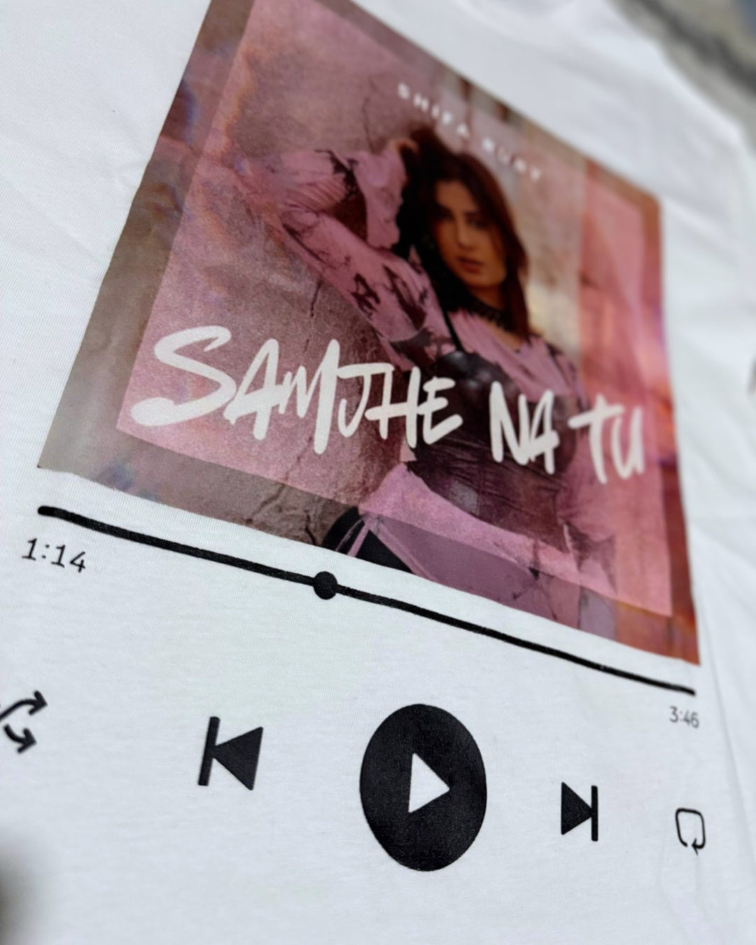 A close-up of a white t-shirt featuring a printed graphic of a music player interface. The graphic includes a pink-toned album cover with the text 'SAMJHE NA TU' in white script, playback controls, and a progress bar.