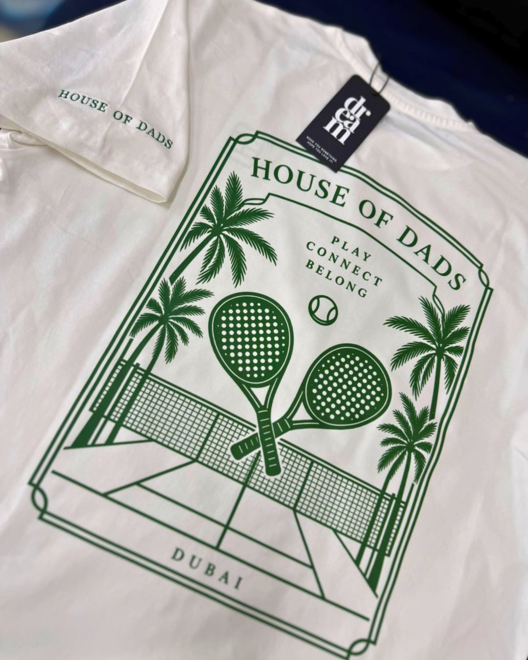 A close-up of a white t-shirt featuring a dark green graphic print on the front. The graphic depicts a tennis court with two crossed tennis rackets, palm trees, and a tennis ball, with the text 'HOUSE OF DADS', 'PLAY CONNECT BELDING', and 'DUBAI'.