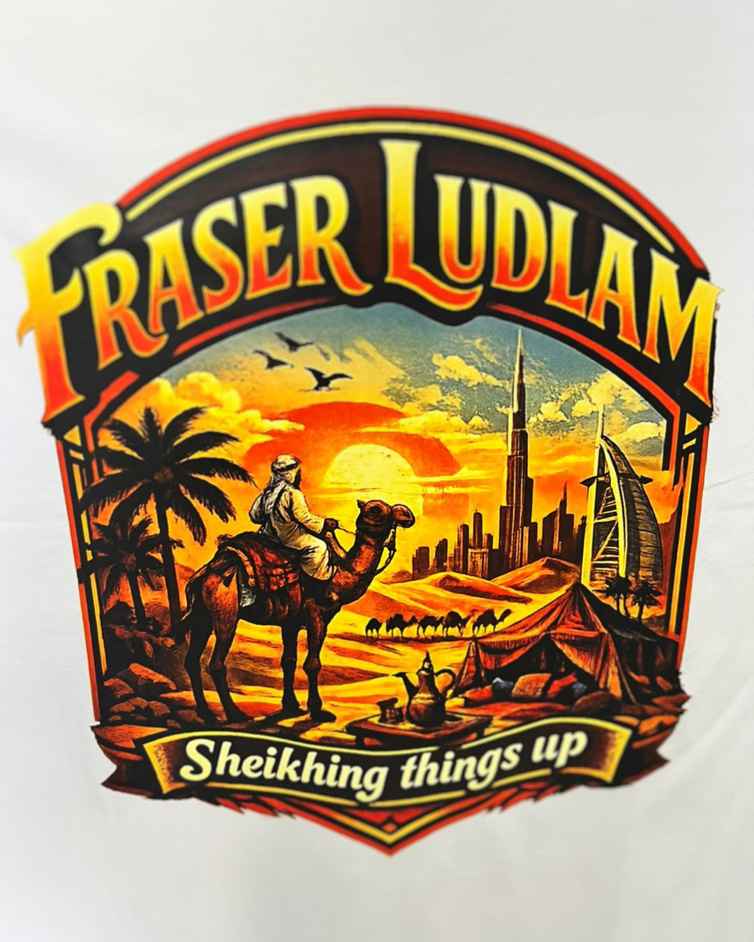 A close-up of a white T-shirt featuring a large graphic print with the text 'FRASER LUDLAM' and 'Sheikhing things up.' The design shows a desert scene with a camel, rider, palm trees, tents, a sunset, and modern skyline elements including the Burj Khalifa and Burj Dubai.
