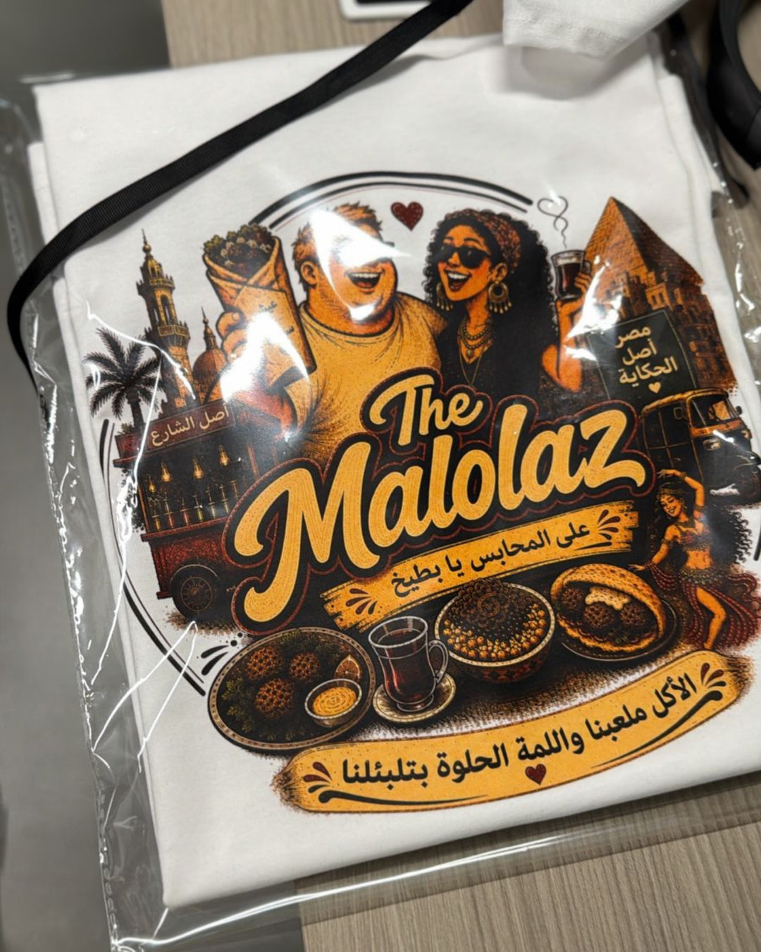 A white t-shirt inside a clear plastic protective sleeve, featuring a large graphic print of 'The Malolaz' coffee shop with Arabic text and illustrations of coffee and pastries.